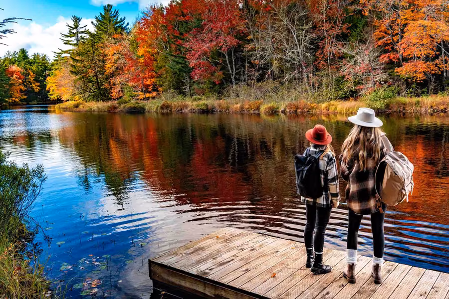 Two hikers on a wooden dock looking across a river lined with autumn colours in Kejimkujik National Park