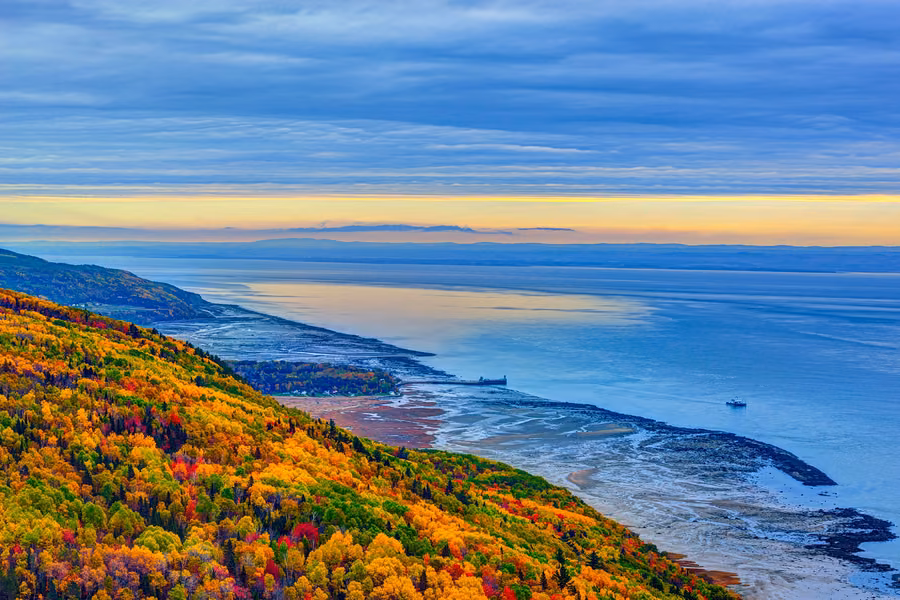 Autumn-coloured hillside sloping down to the St Lawrence River's tidal flats and a long wharf in Charlevoix