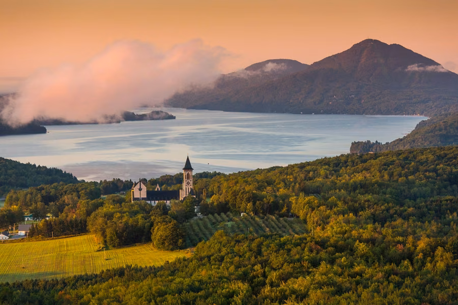 Abbaye de Saint-Benoît-du-Lac among farmland and forest above Lake Memphremagog, with misty mountains at sunrise