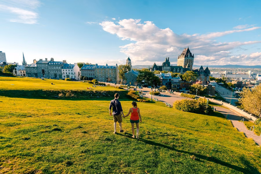 Couple walking across a sunlit grassy slope towards Old Québec's rooftops and the Château Frontenac