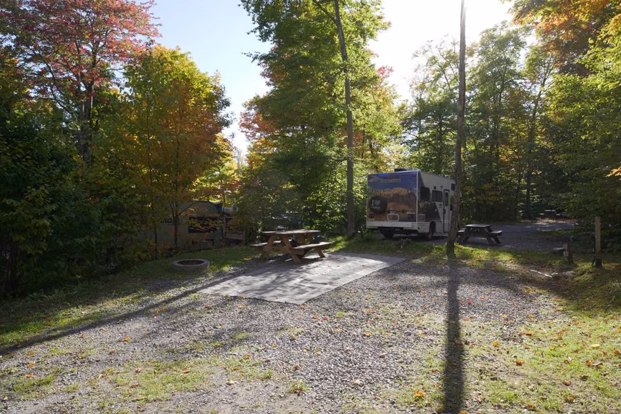 Wooded campsite with a gravel pad, picnic tables and a fire ring, with a motorhome parked among autumn trees at Camping de la Joie