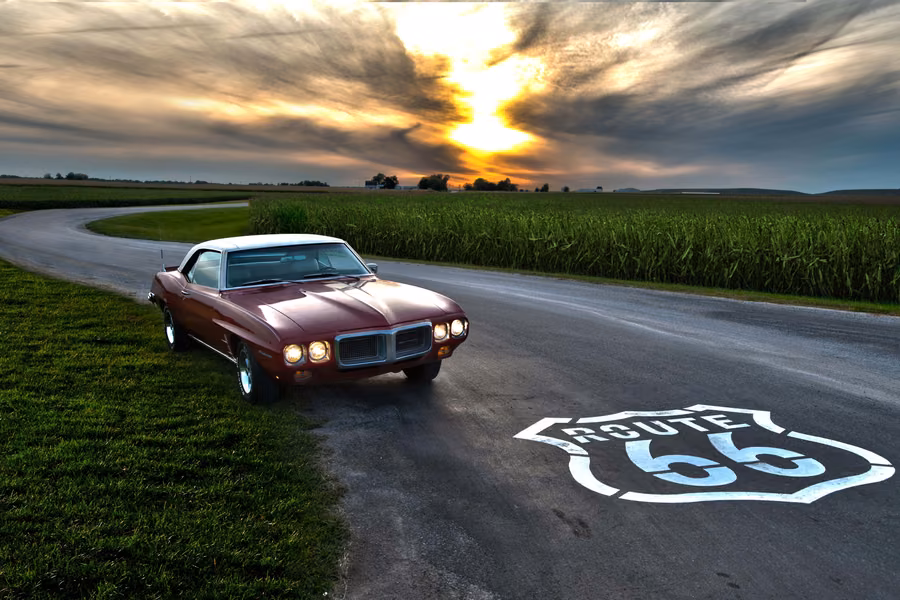 Classic red Pontiac Firebird on a country road beside a cornfield at sunset, with a Route 66 shield painted on the road