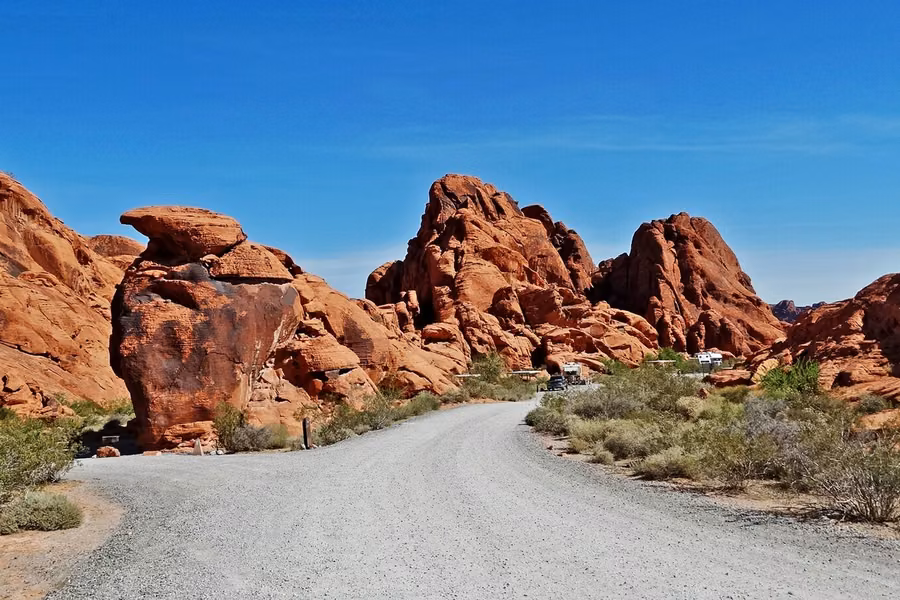 Gravel campground road winding between red sandstone outcrops and a balanced rock, with RVs parked in the distance, at Valley of Fire State Park
