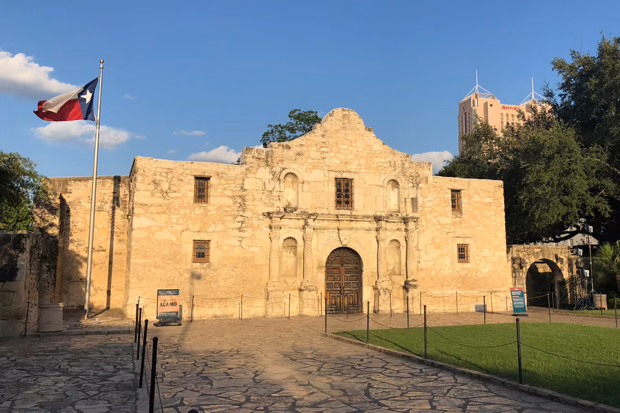 The Alamo's carved limestone church front under a blue sky, with the Texas flag flying on Alamo Plaza in San Antonio