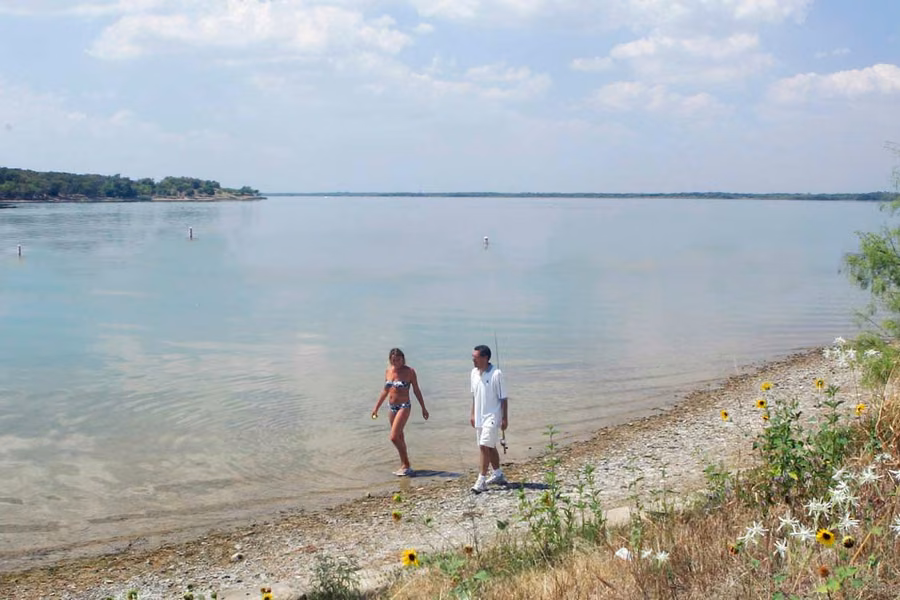 Couple walking along the pebbly shore of Joe Pool Lake at Cedar Hill State Park, with sunflowers on the bank