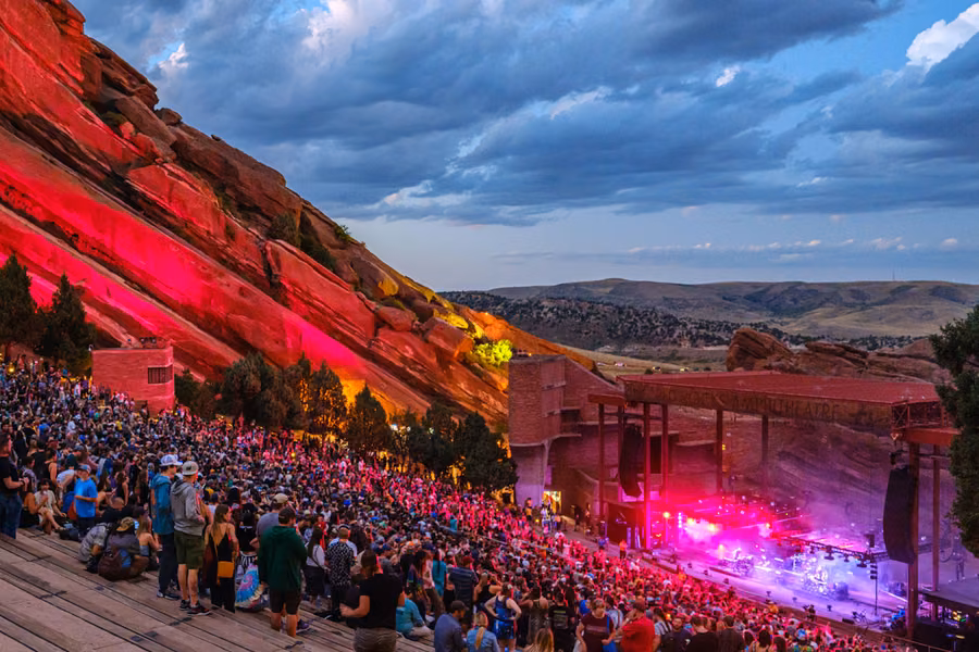 Crowd filling the Red Rocks Amphitheatre at dusk, with the sandstone walls lit red and the stage glowing below