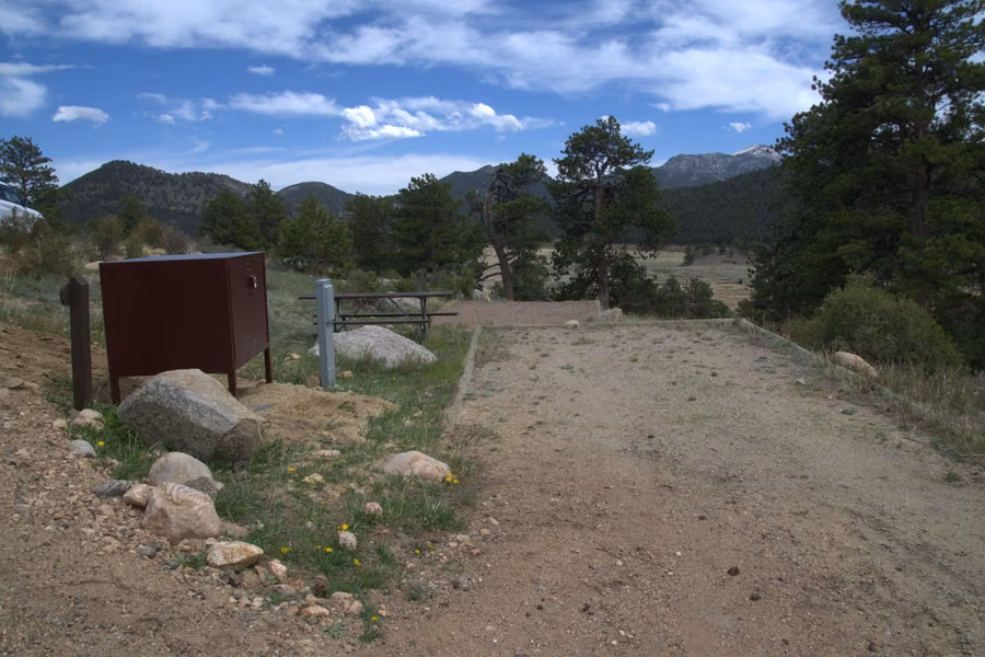 Empty campsite with a brown food storage box and picnic table among pines at Moraine Park Campground, with forested mountains behind