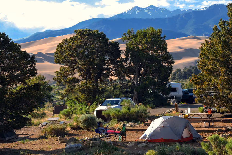 Tent and campsite among piñon pines at Piñon Flats Campground, with the Great Sand Dunes and snow-capped peaks behind