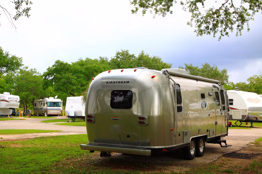 Silver Airstream caravan parked on a grassy RV site among shade trees, with other caravans and a motorhome behind, at Larry and Penny Thompson Park