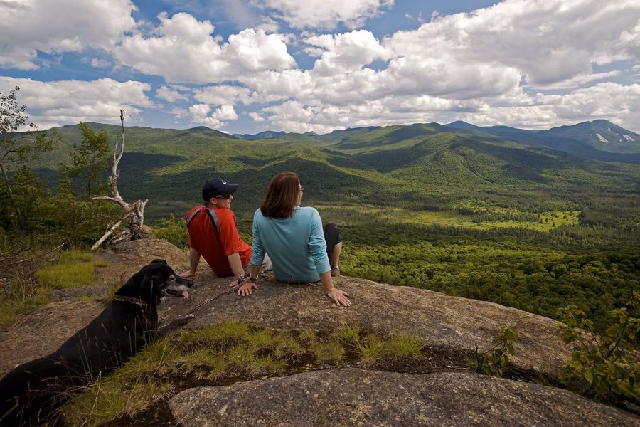 Two hikers and a black dog resting on a rocky summit above forested Adirondack mountains and valleys