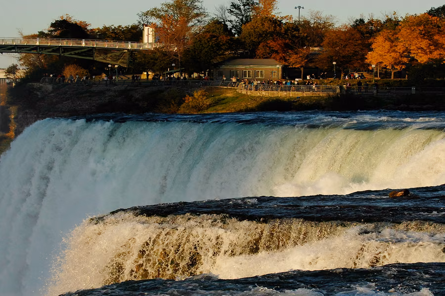 White water pouring over the brink of the falls at Niagara Falls State Park, with a bridge and autumn trees behind