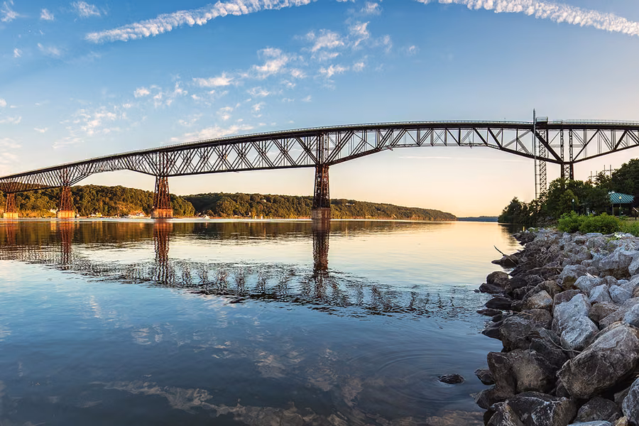 The long steel truss of the Walkway Over the Hudson reflected in the calm Hudson River under a blue sky
