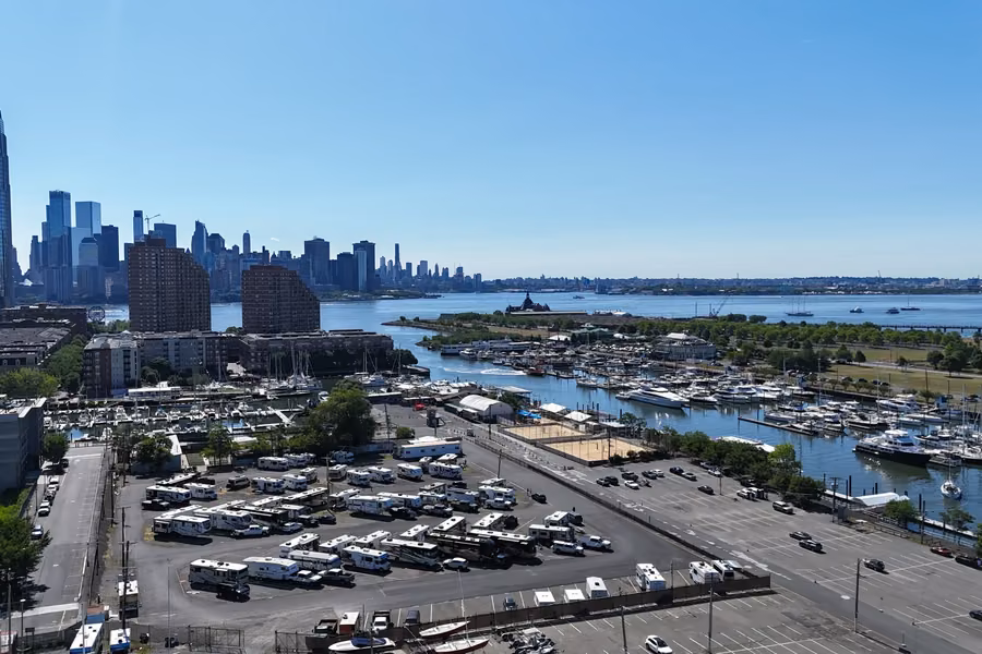RVs parked on the paved lot at Liberty Harbor RV Park beside a marina, with Jersey City towers and the harbour behind