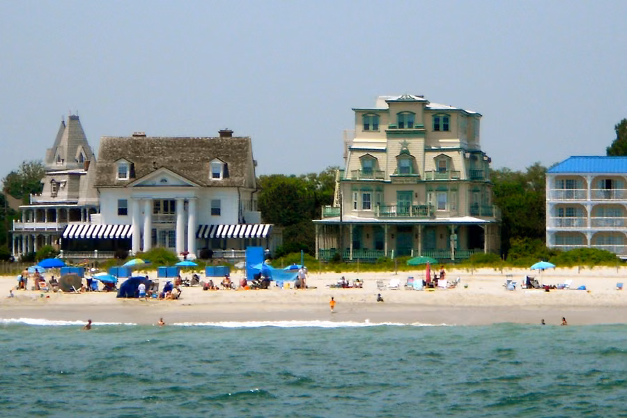 Victorian houses lining Beach Avenue in Cape May behind a sandy beach with umbrellas and sunbathers, seen from the sea