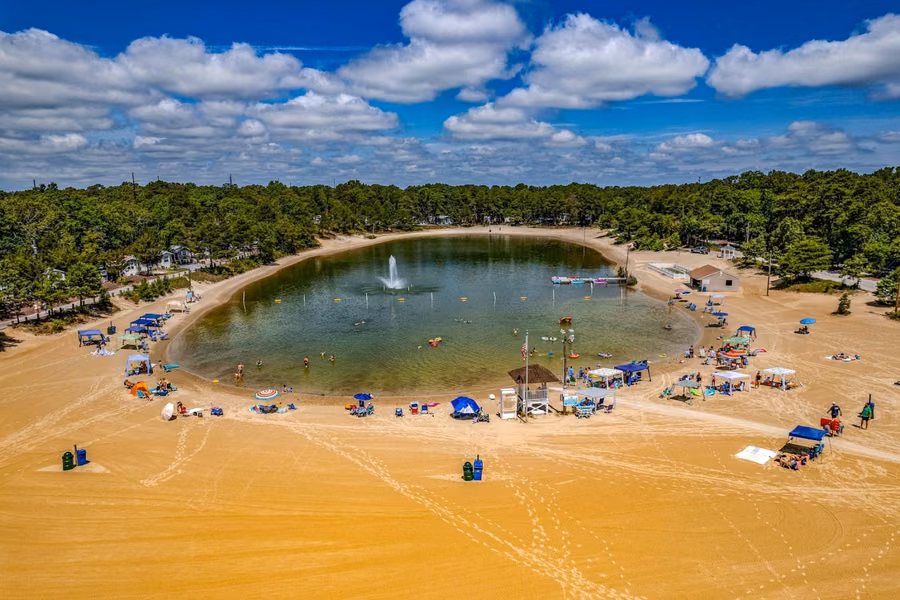 Aerial view of the freshwater lake with a fountain and wide sandy beach at Ocean View Resort Campground, ringed by pine forest