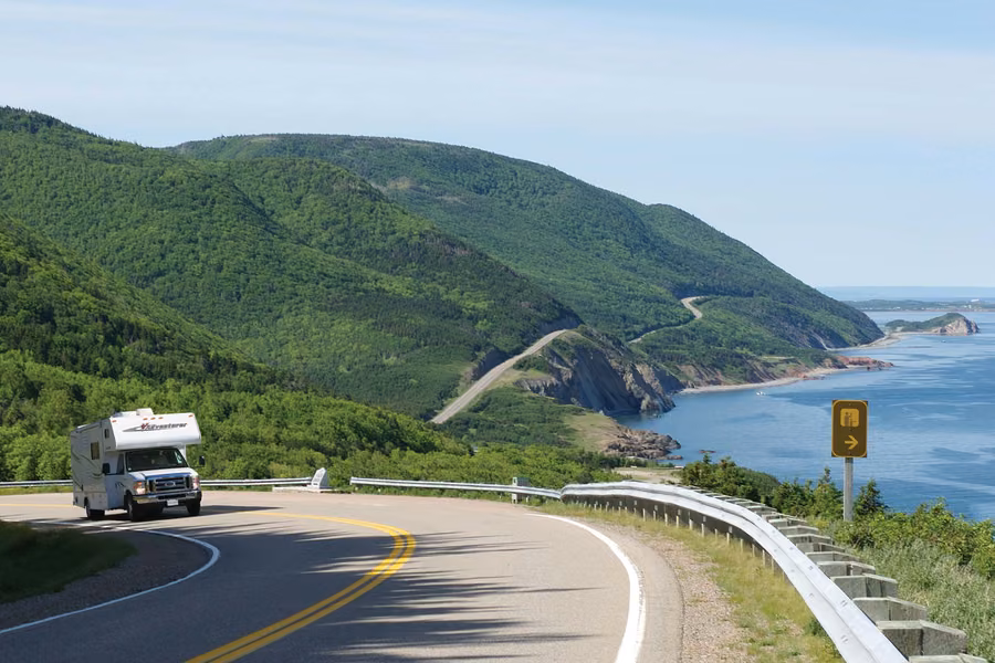 Motorhome rounding a bend on the Cabot Trail, with green highlands and the road winding along the ocean coast behind