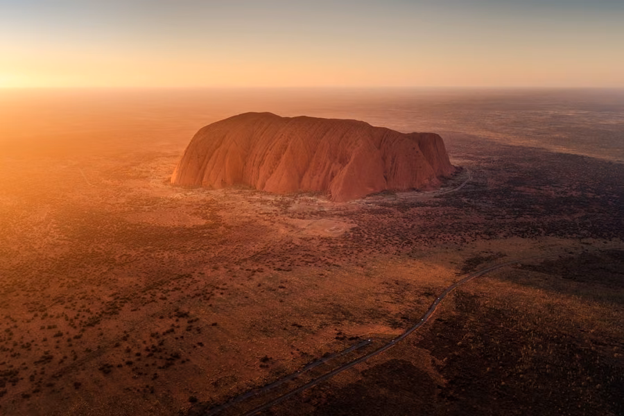 Aerial view of Uluṟu glowing in the sunrise light above red desert plains in Uluṟu-Kata Tjuṯa National Park