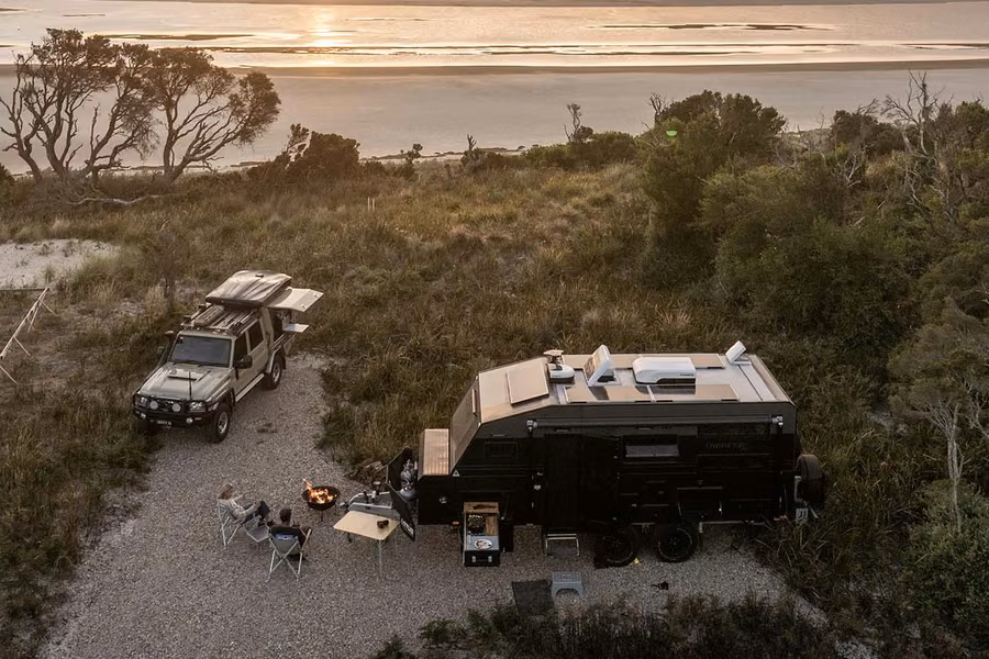 Campers relaxing beside a 4WD and an off-road caravan on a bush campsite above a tidal bay at sunset in the Bay of Fires area