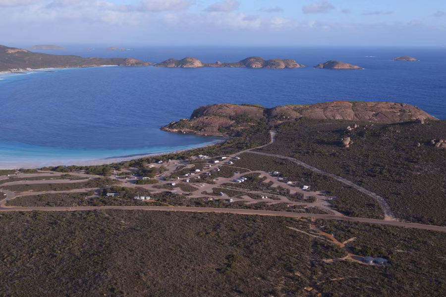 Aerial view of Lucky Bay Campground in the heath beside a white beach and blue bay, with granite headlands and islands offshore