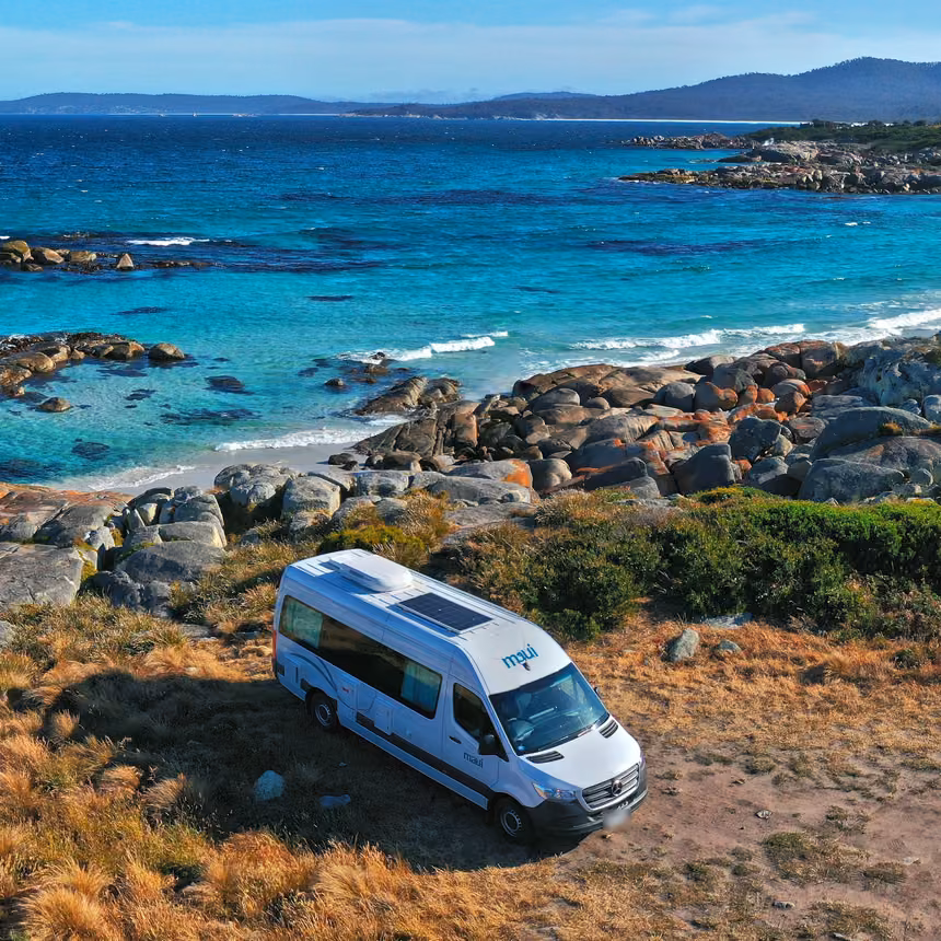 maui campervan parked on a grassy headland above granite boulders, turquoise water and a white sand beach on the Tasmanian coast
