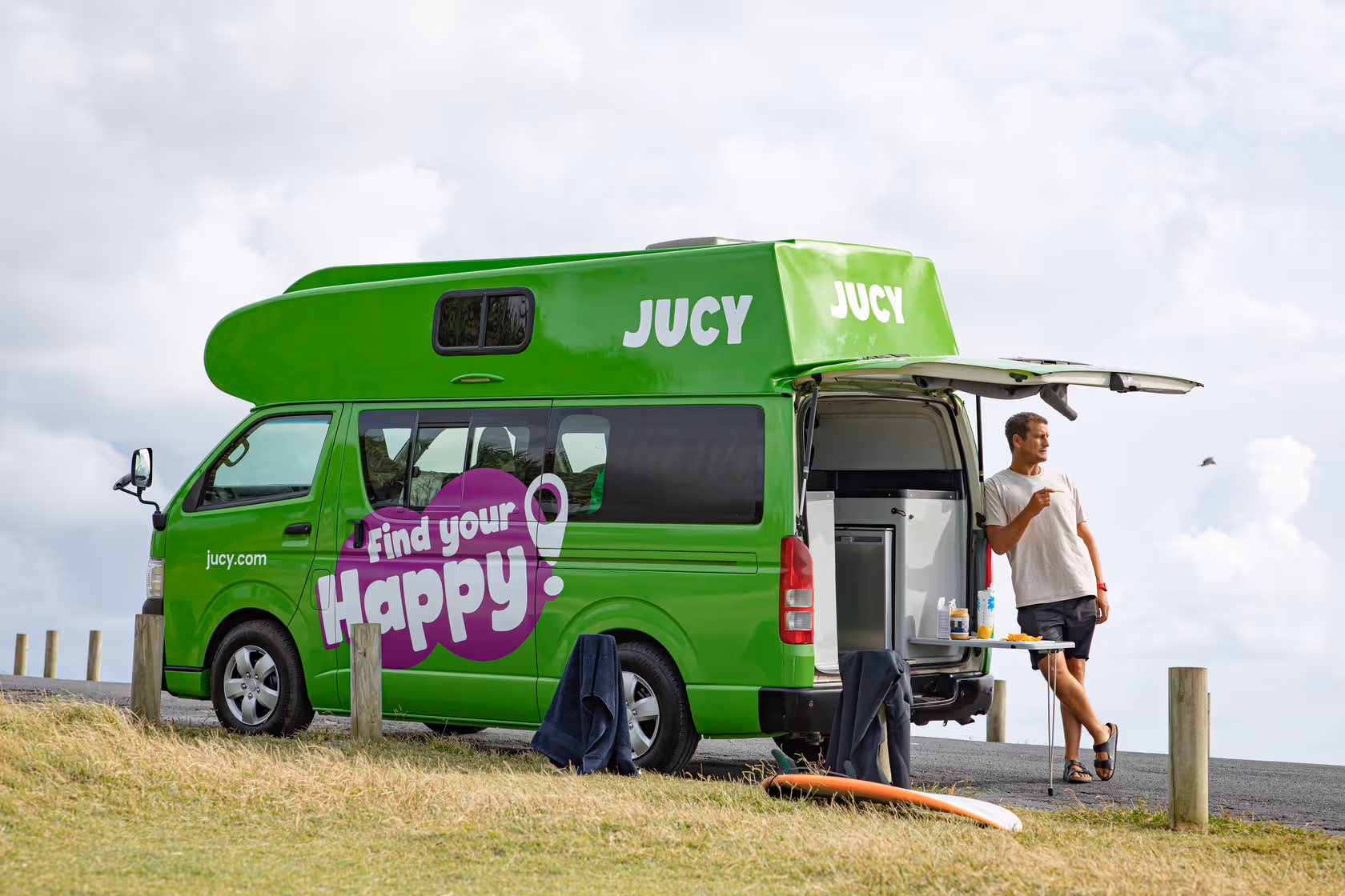 Green JUCY campervan parked roadside with open rear, traveler at small table