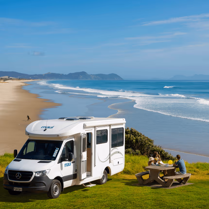 Maui campervan parked on a grassy clifftop overlooking a stunning New Zealand beach, with travellers enjoying a picnic by the ocean — showcasing campervan hire NZ, coastal road trips, and scenic motorhome adventures.