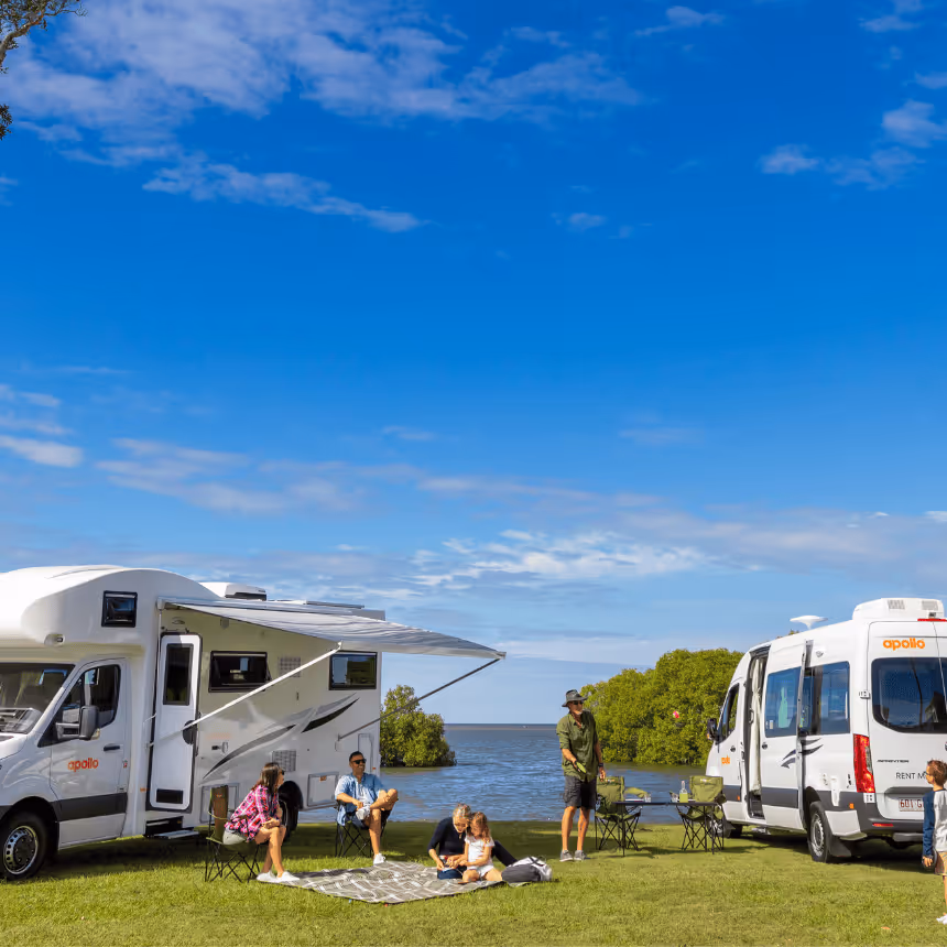 Families enjoying a lakeside picnic beside Apollo campervans on a sunny day in Australia. Two modern motorhomes are parked on green grass near the water, showcasing Apollo’s campervan rentals — perfect for family road trips, outdoor holidays, and Australia