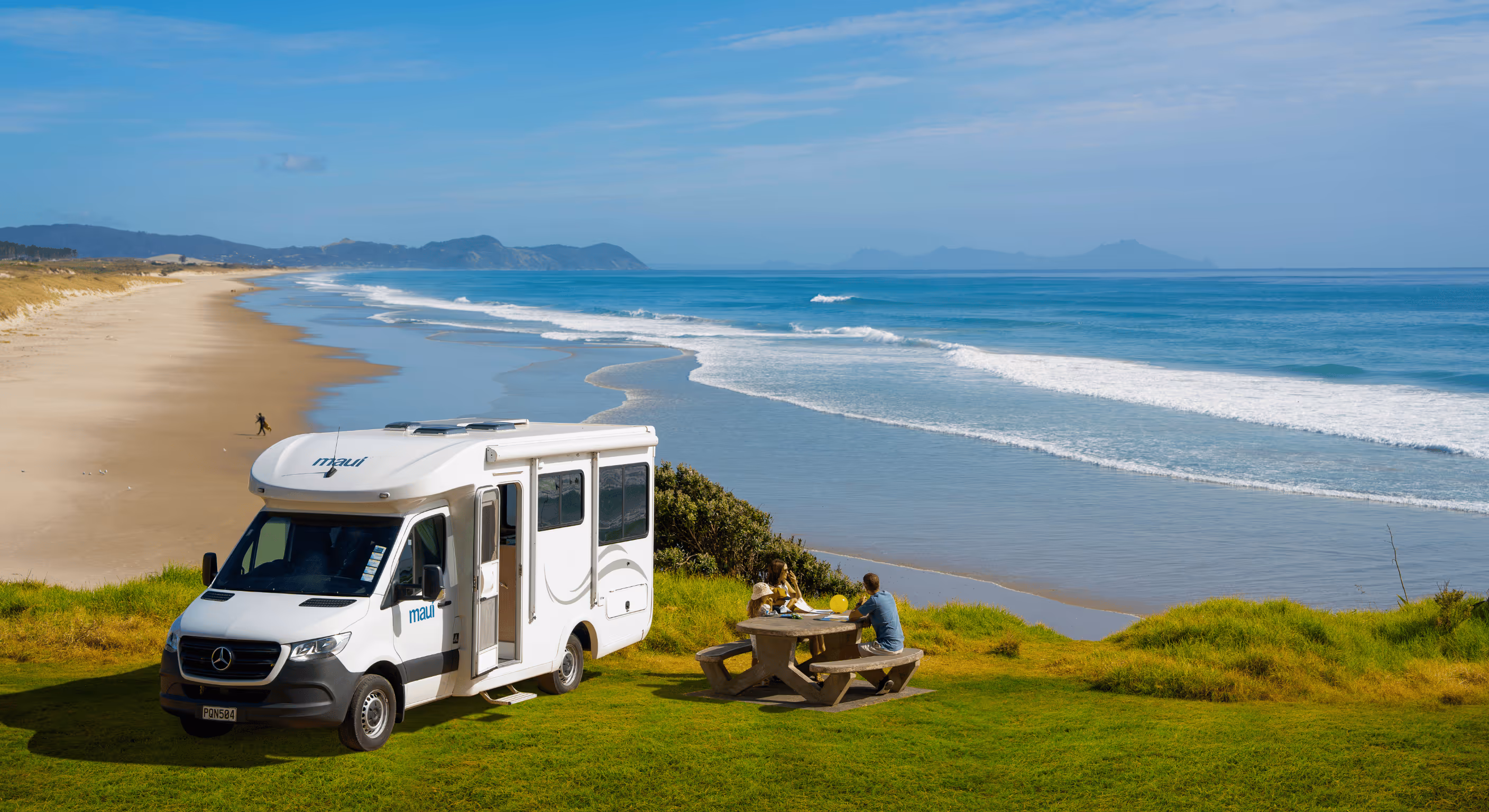 Maui campervan parked on a grassy clifftop overlooking a stunning New Zealand beach, with travellers enjoying a picnic by the ocean — showcasing campervan hire NZ, coastal road trips, and scenic motorhome adventures.