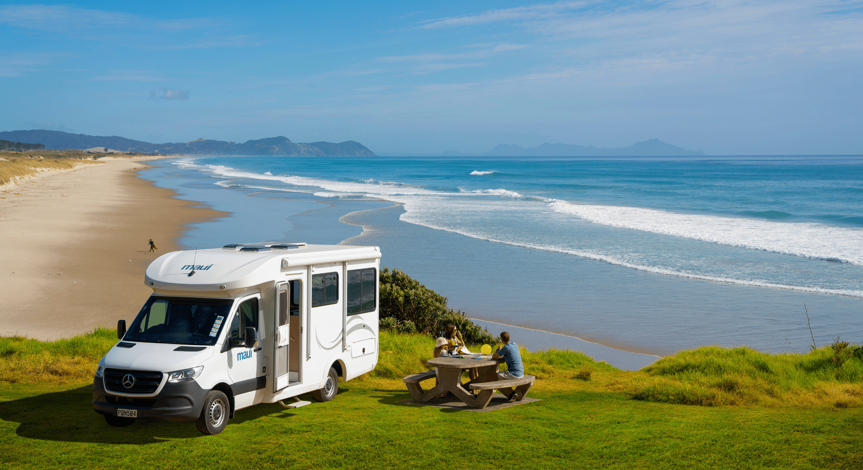 Maui campervan parked on a grassy clifftop overlooking a stunning New Zealand beach, with travellers enjoying a picnic by the ocean — showcasing campervan hire NZ, coastal road trips, and scenic motorhome adventures.