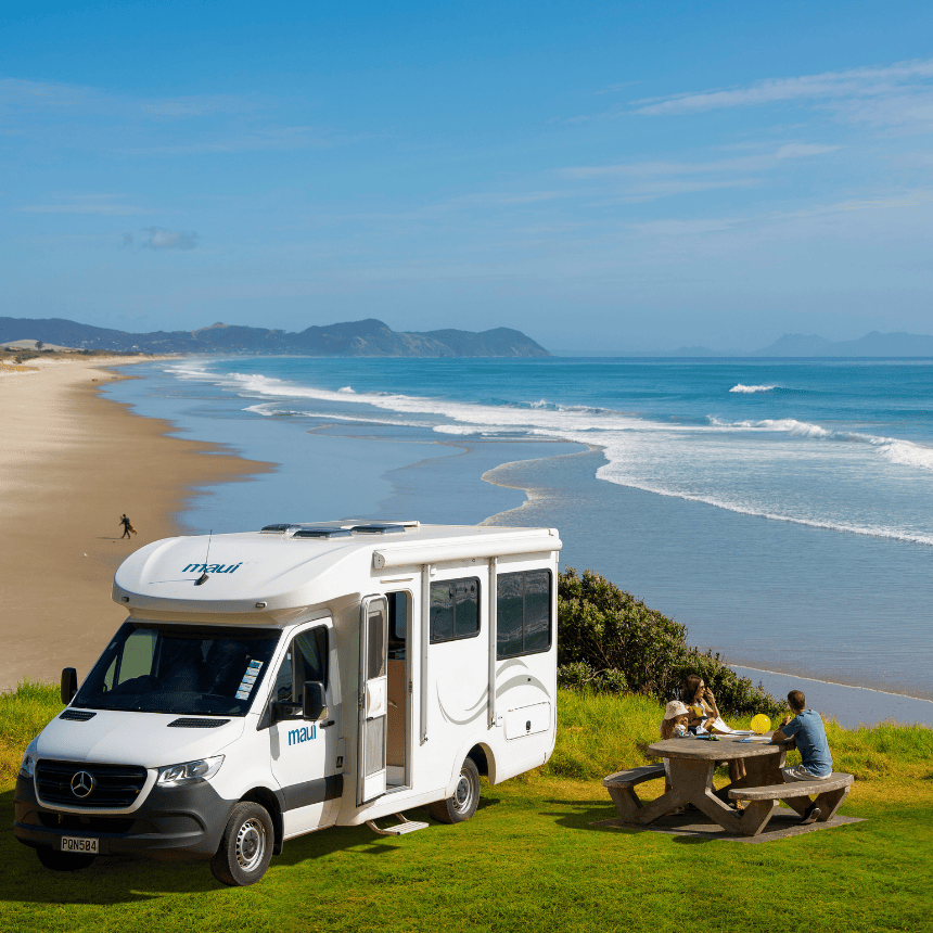 Maui campervan parked on a grassy clifftop overlooking a stunning New Zealand beach, with travellers enjoying a picnic by the ocean — showcasing campervan hire NZ, coastal road trips, and scenic motorhome adventures.