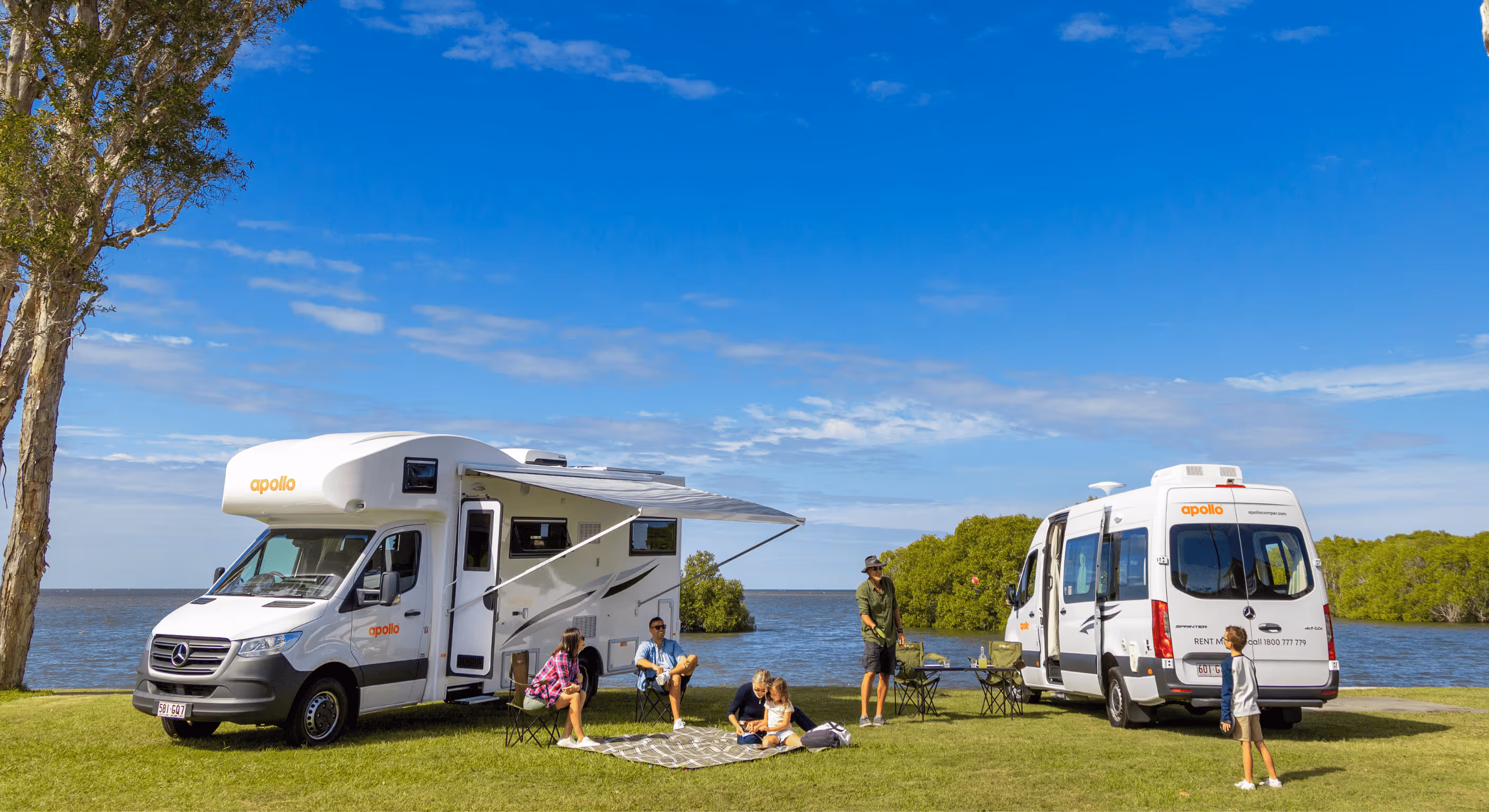 Families enjoying a lakeside picnic beside Apollo campervans on a sunny day in Australia. Two modern motorhomes are parked on green grass near the water, showcasing Apollo’s campervan rentals — perfect for family road trips, outdoor holidays, and Australia
