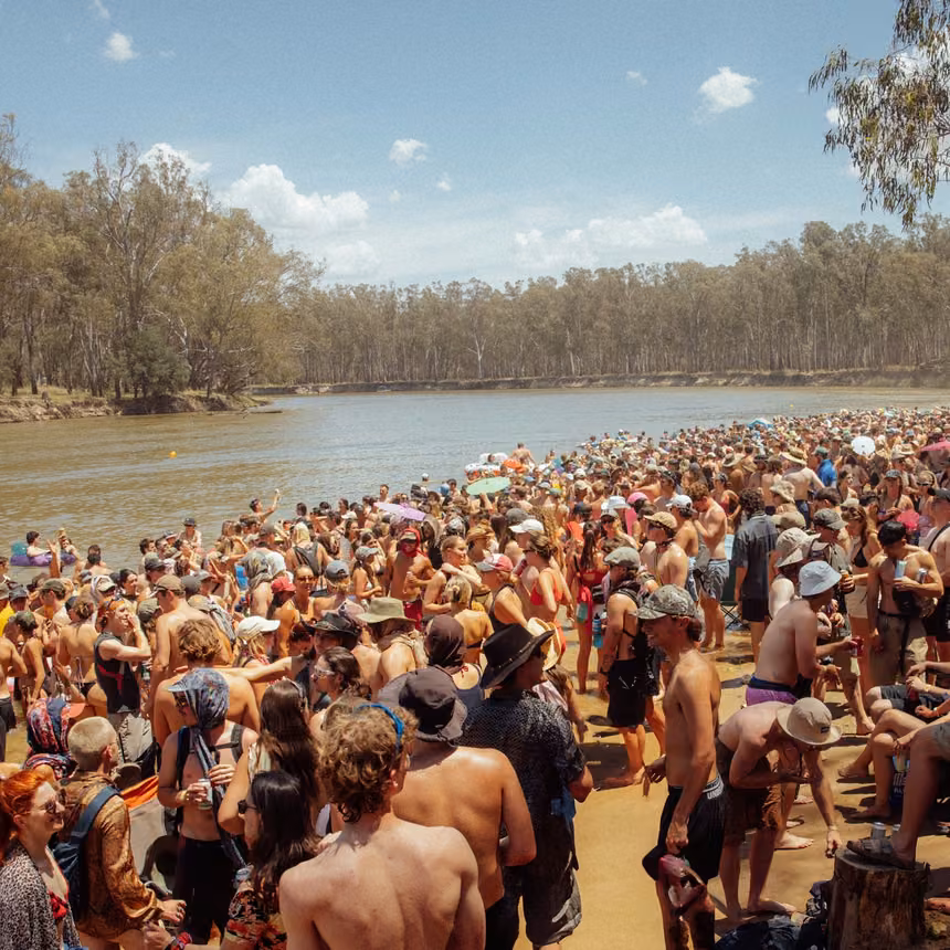 A large festival crowd on a sandy beach beside the Murray River, with red gum forest on the far bank, at Strawberry Fields