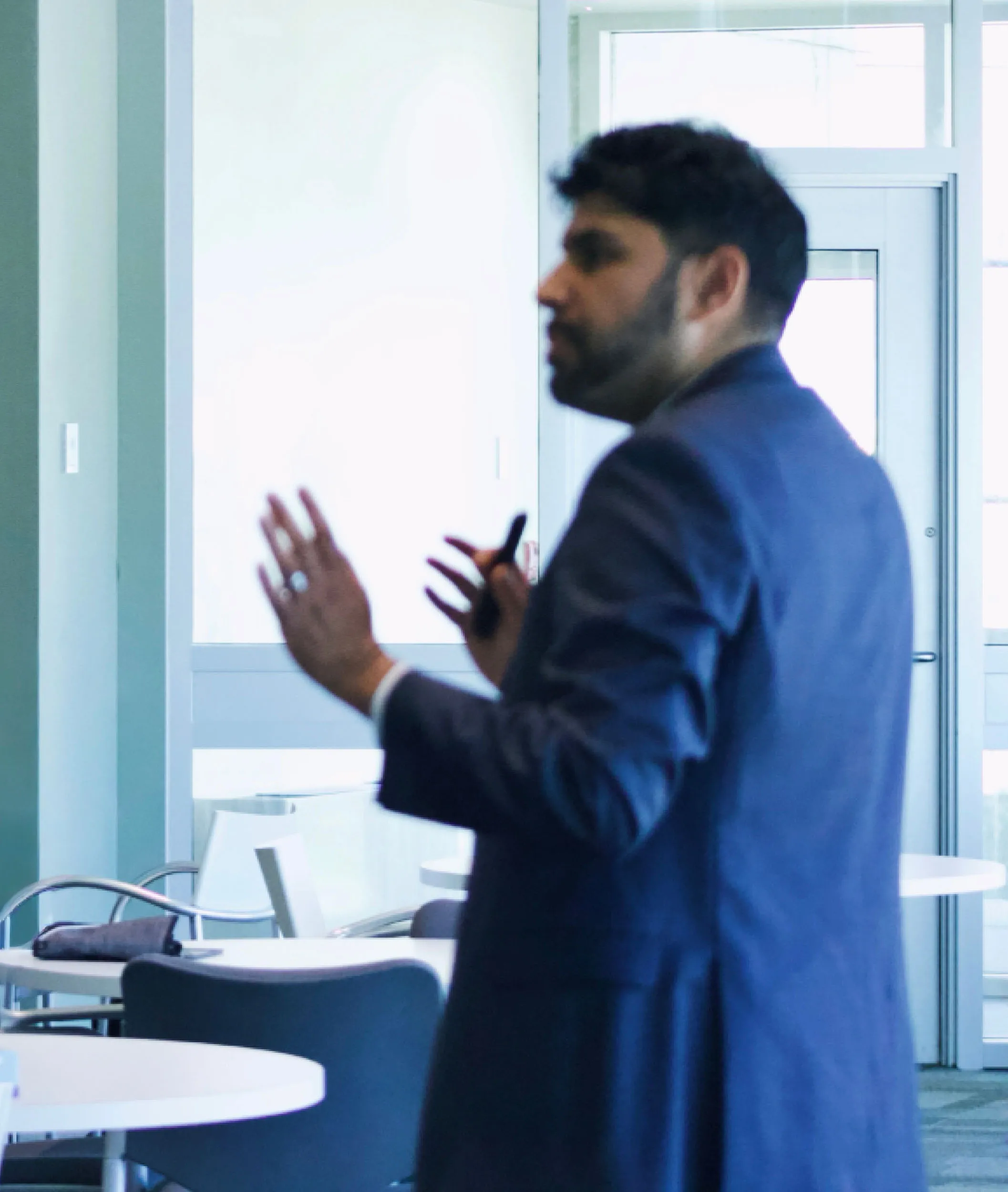 Man in a navy blazer giving a presentation in a bright room with tables and chairs.