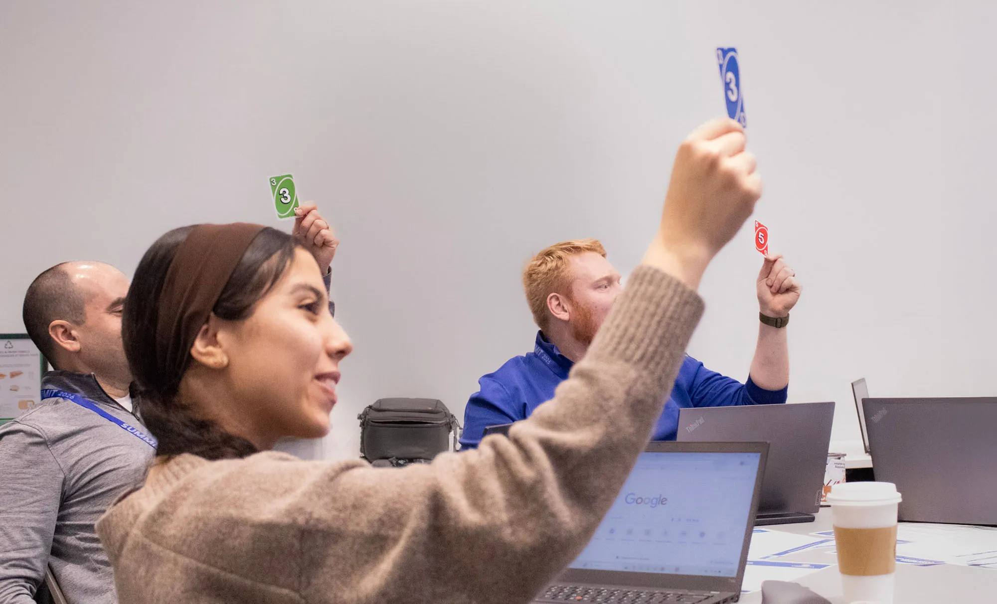Three people sitting at a table with laptops, raising Uno cards during a group activity or meeting.
