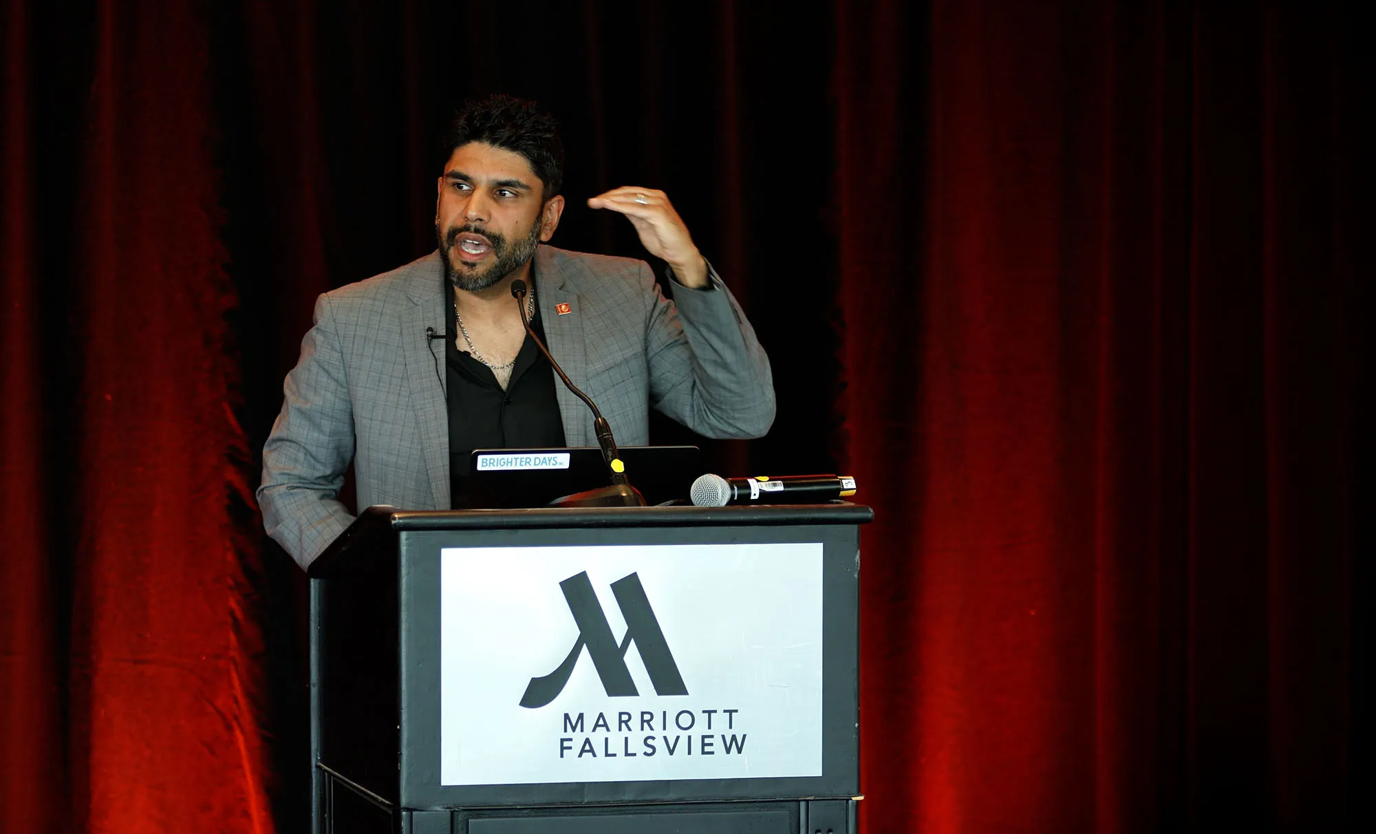 Man in a grey suit jacket speaking at a Marriott Fallsview podium with a microphone, gesturing with his right hand.