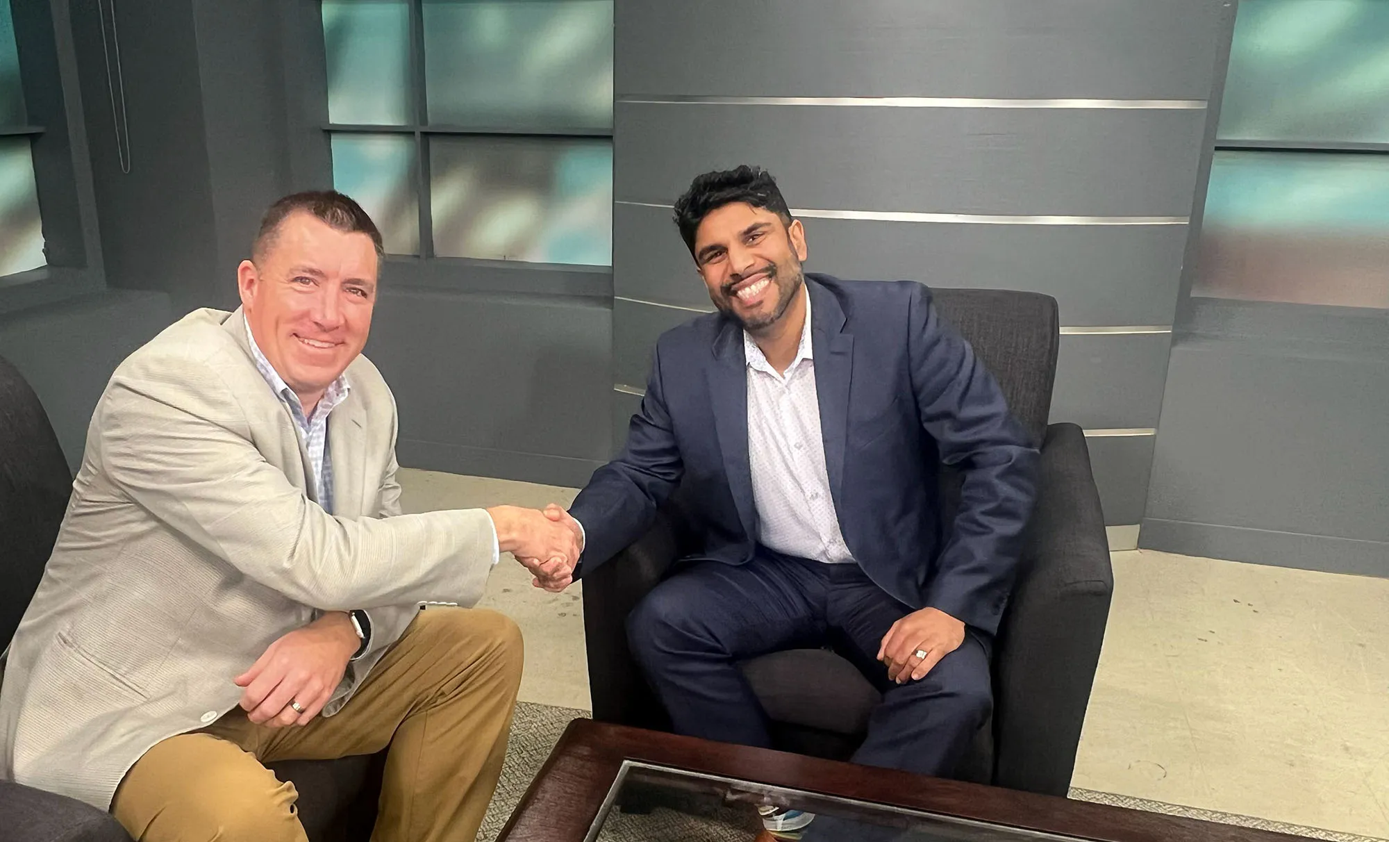 Two men sitting in an office shaking hands and smiling at the camera.