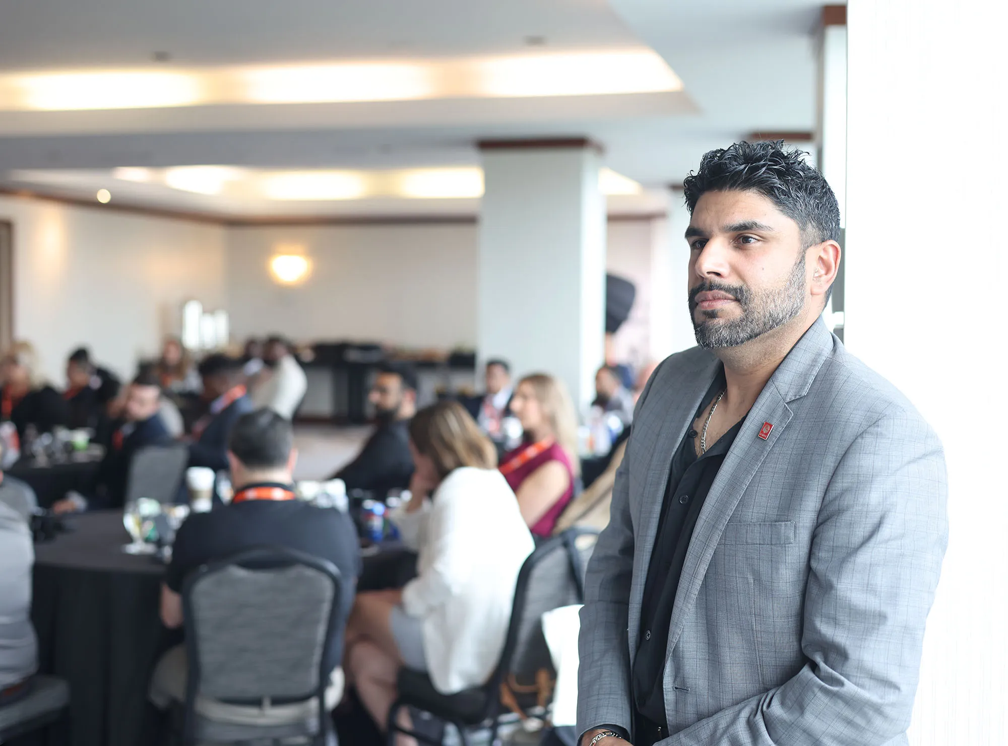 Man in gray blazer standing thoughtfully by a window with a blurred group of people seated at tables in a conference room.