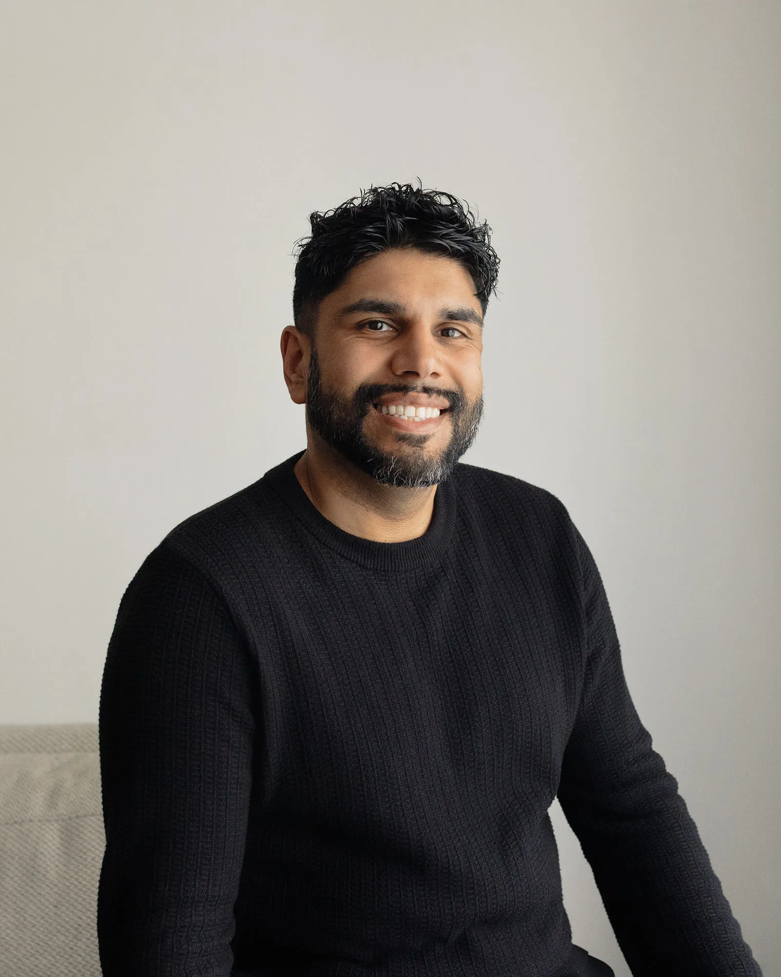 Smiling man with dark hair and beard wearing a black sweater sitting against a light background.