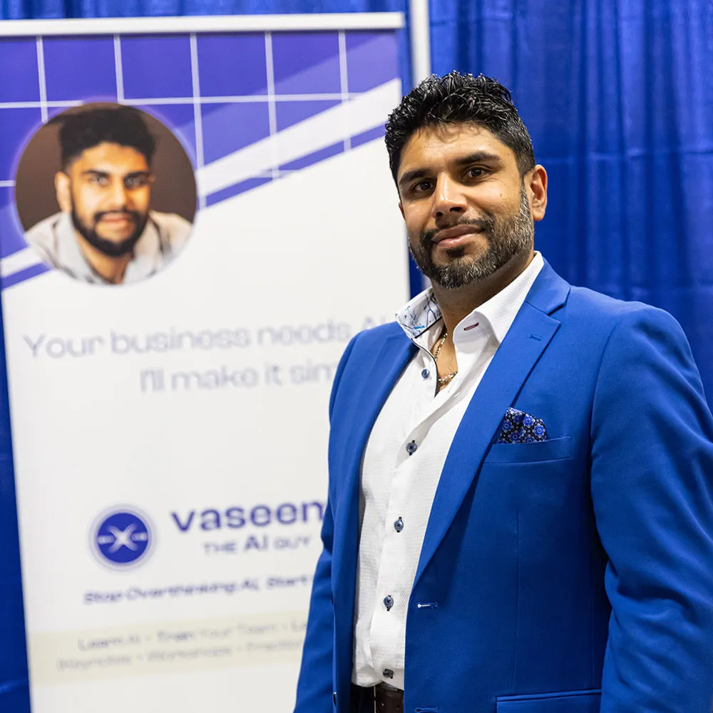 Man with dark hair and beard wearing a blue blazer and white shirt standing in front of a blue and white promotional banner.
