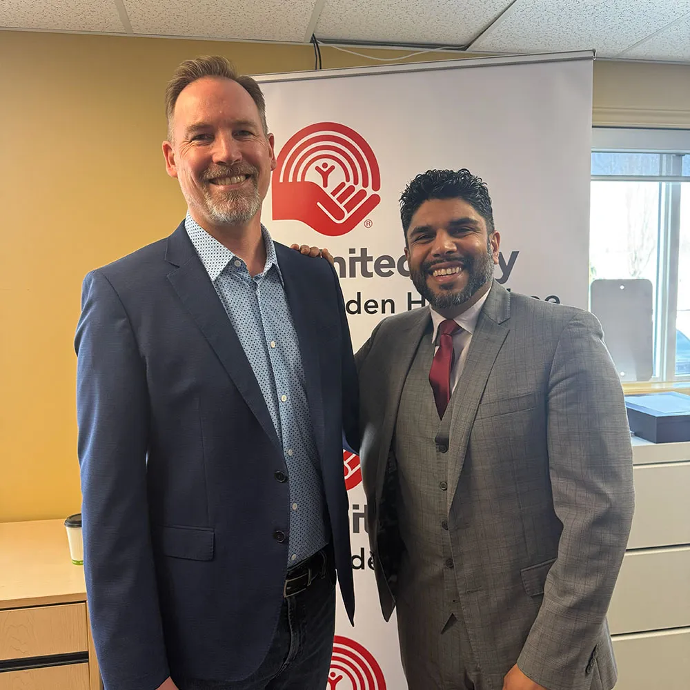 Two men in suits smiling in front of a United Way banner inside an office.