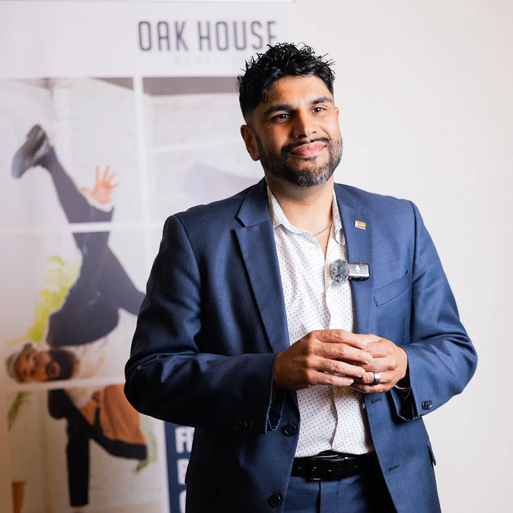 Smiling man with beard wearing a blue suit and white polka dot shirt, standing indoors with a blurred background banner reading 'Oak House'.