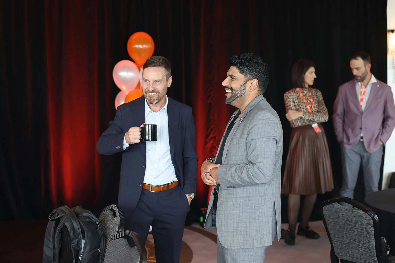 Two men in business attire smiling and conversing at a professional event with balloons in the background, while a woman and another man talk in the background.