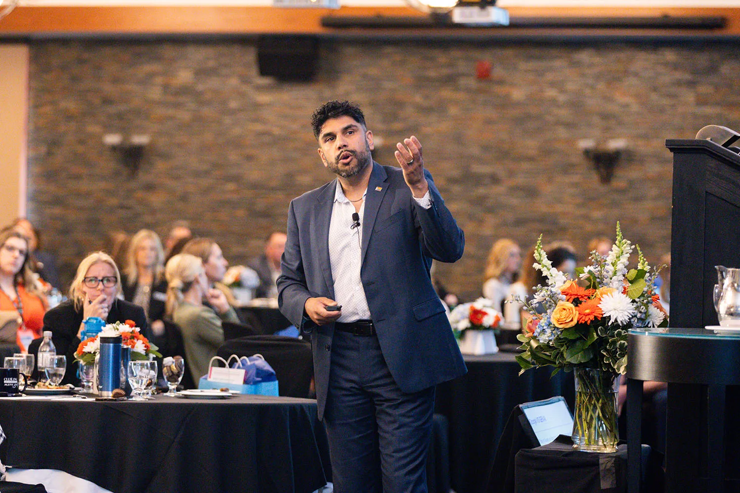 Man in a blue suit giving a speech or presentation to an attentive audience in an indoor conference setting.