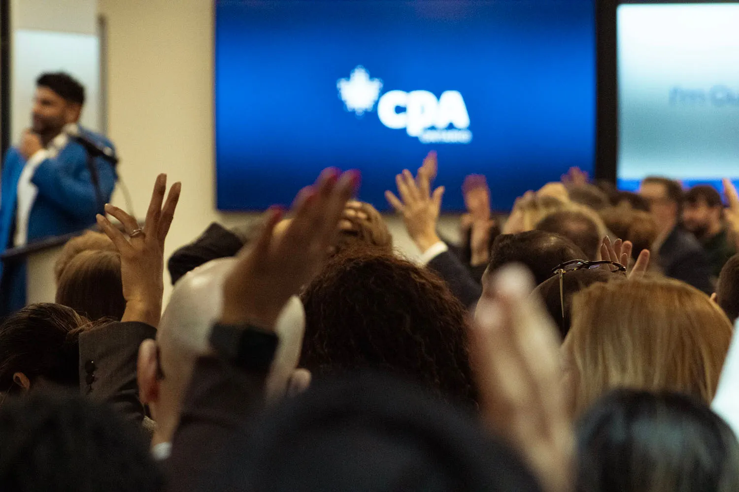 Audience raising hands during a presentation with a speaker and a blue screen displaying the CPA logo in the background.