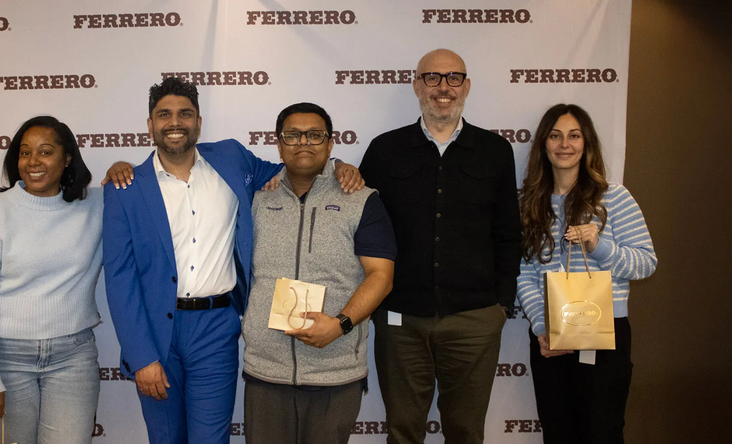 Six people smiling and standing together in front of a Ferrero branded backdrop, some holding gift bags.
