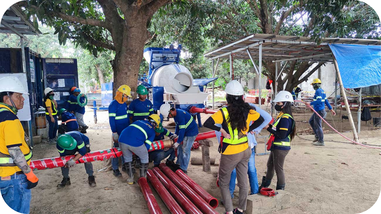 A large, diverse construction crew in hard hats works together to assemble long sections of a red concrete pump pipe outdoors.