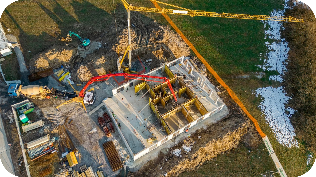 A high-altitude aerial view of a new house construction, showing a crane and a concrete pump pouring the basement walls.