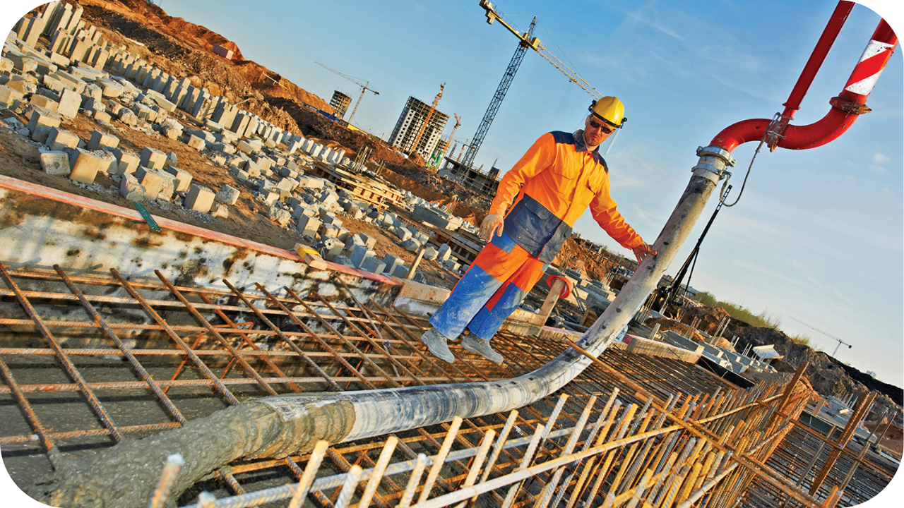 From a low angle, a worker in a hard hat guides a large pump hose, pouring concrete into a rebar-lined foundation trench.