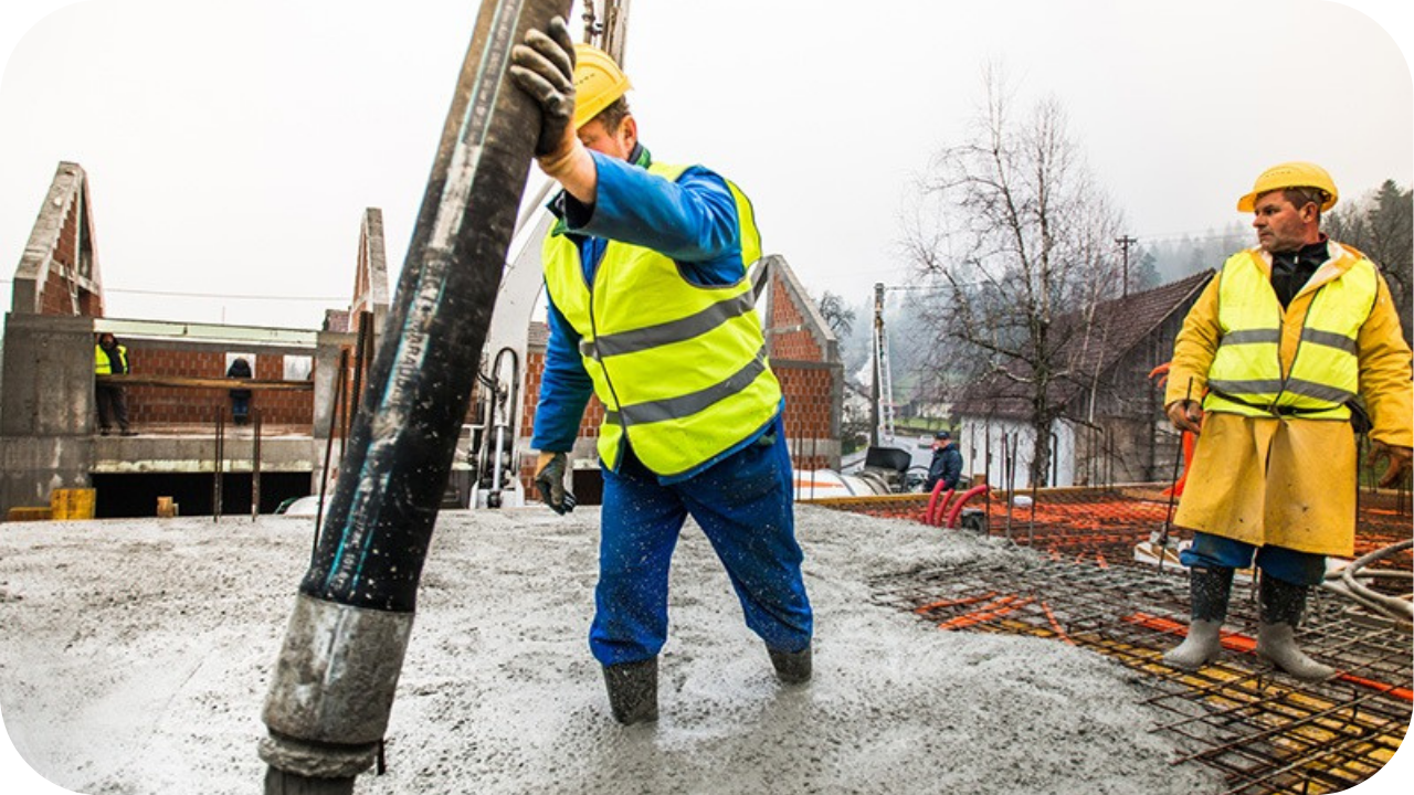 Two workers in hard hats stand in wet concrete on a suspended slab, with one guiding the large pump hose during a pour.