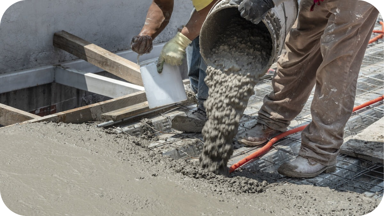 Construction workers pour wet concrete from large white buckets onto a steel rebar grid to create a new slab.